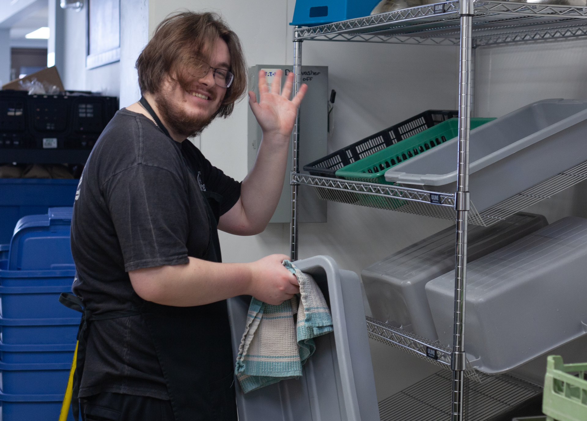TGP_Volunteers_V2 Volunteer waving at the camera while drying bins in the Meal Centre kitchen.